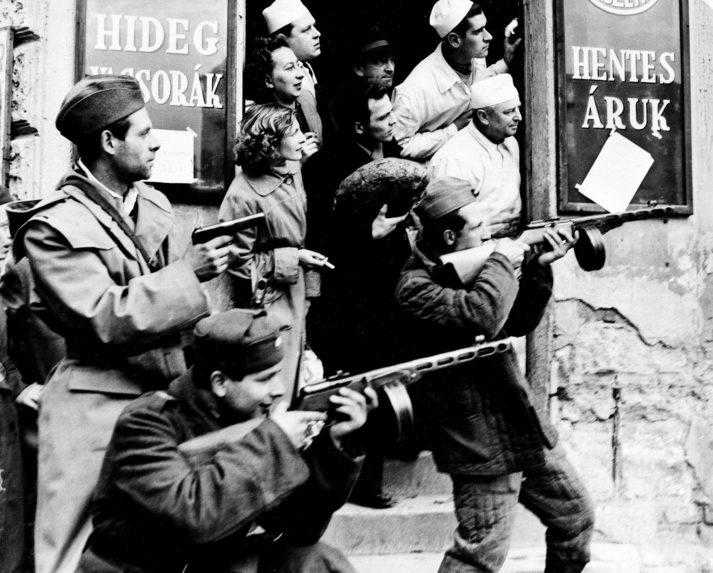 Zákazníci a zamestnanci miestnej predajne sa pozerajú na maďarské revolučné sily, ktoré mieria na príslušníkov tajnej komunistickej polície.