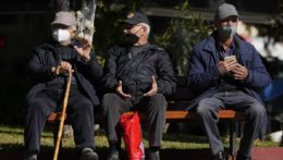 traja starší muži sedia s ochrannými rúškami na lavičke v gréckych Aténach