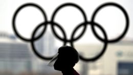 Osoba s ochranným štítom na tvári kráča okolo olympijských kruhov.