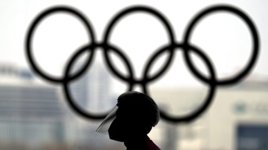 Osoba s ochranným štítom na tvári kráča okolo olympijských kruhov.