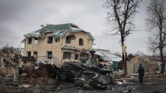 Muž sa pozerá na zničenú budovu a ruské vojenské vozidlá na ceste v meste Bucha.