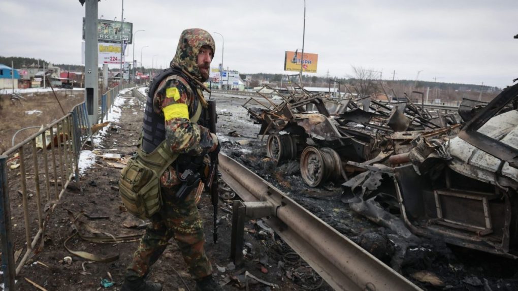 Ozbrojený muž stojí vedľa vrakov ruských vojenských vozidiel v meste Buča.
