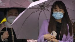Ľudia s ochrannými rúškami kráčajú cez križovatku v daždivom ráne v čínskom Pekingu.
