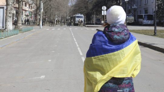 žena s ukrajinskou vlajkou stojí pred ruskými vojakmi počas protestu proti okupácii v Chersone