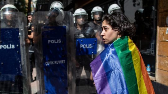 Žena s dúhovou vlajkou stojí pred blokádou policajtov v tureckom Istanbule.