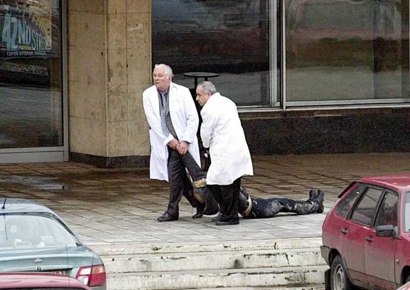 Na snímke dvaja muži v lekárskych plášťoch ťahajú po zemi telo tretej osoby.