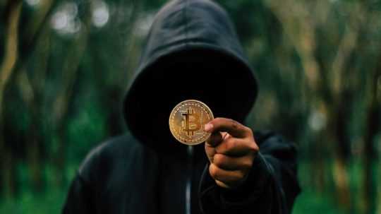 Postava v kapucni drží mincu Bitcoinu.