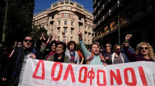 Demonštranti držia v rukách plagát s nápisom "vrahovia" v gréčtine.