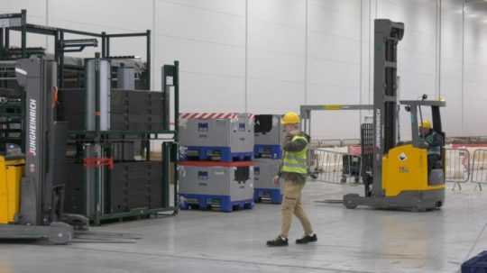 Na snímke pracovník vo výrobnej hale s helmou na hlave a okolo neho dva vysokozdvižné vozíky.