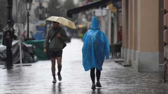 Na snímke je muž s dáždnikom a osoba v pršiplášti na upršanej ulici.