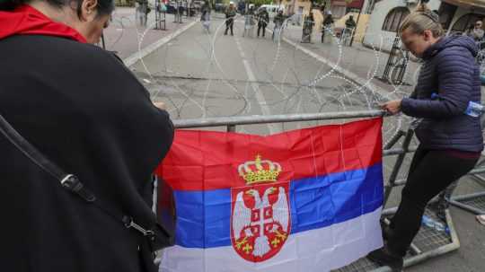 Ženy upevňujú srbskú zástavu na oplotenie počas protestu.