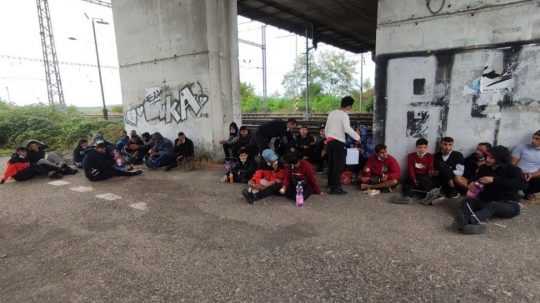 na snímke skupinka migrantov sediaca na zemi pod mostom.