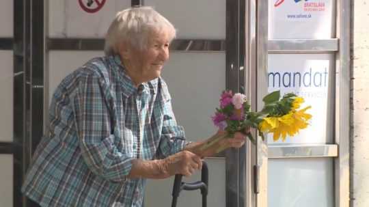 Na snímke je dôchodkyňa opretá o rukoväť nákupnej tašky s kvetmi v ruke.