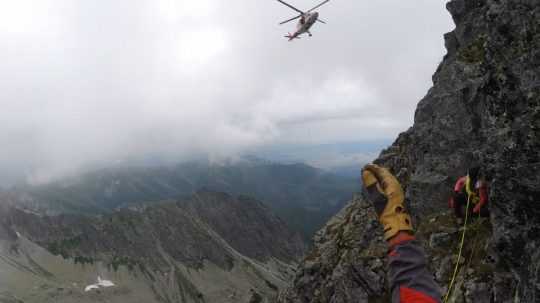 Na snímke je časť hory, ruka záchranára a prelietajúci vrtuľník.