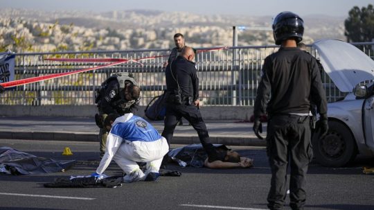 Izraelský policajt mieri na muža pištoľou, keď záchranári prehľadávajú miesto streľby v Jeruzaleme.
