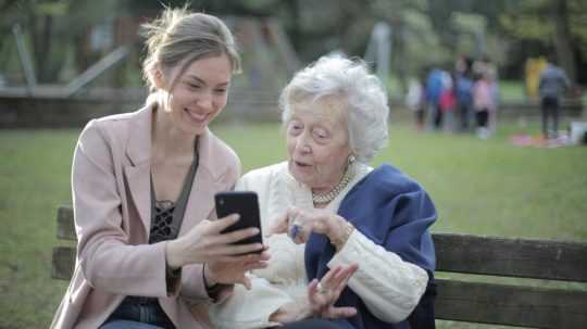 Na snímke vľavo mladá žena ukazuje staršej pani vpravo mobilný telefón. Scéna sa odohráva na lavičke v parku.