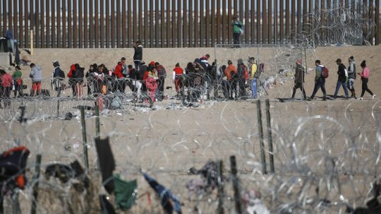 Migranti stoja v rade pri hraničnom múre USA po zadržaní americkými imigračnými úradmi.