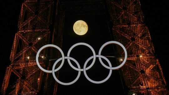 Spln mesiaca vychádza za olympijskými kruhmi visiacimi z Eiffelovej veže.